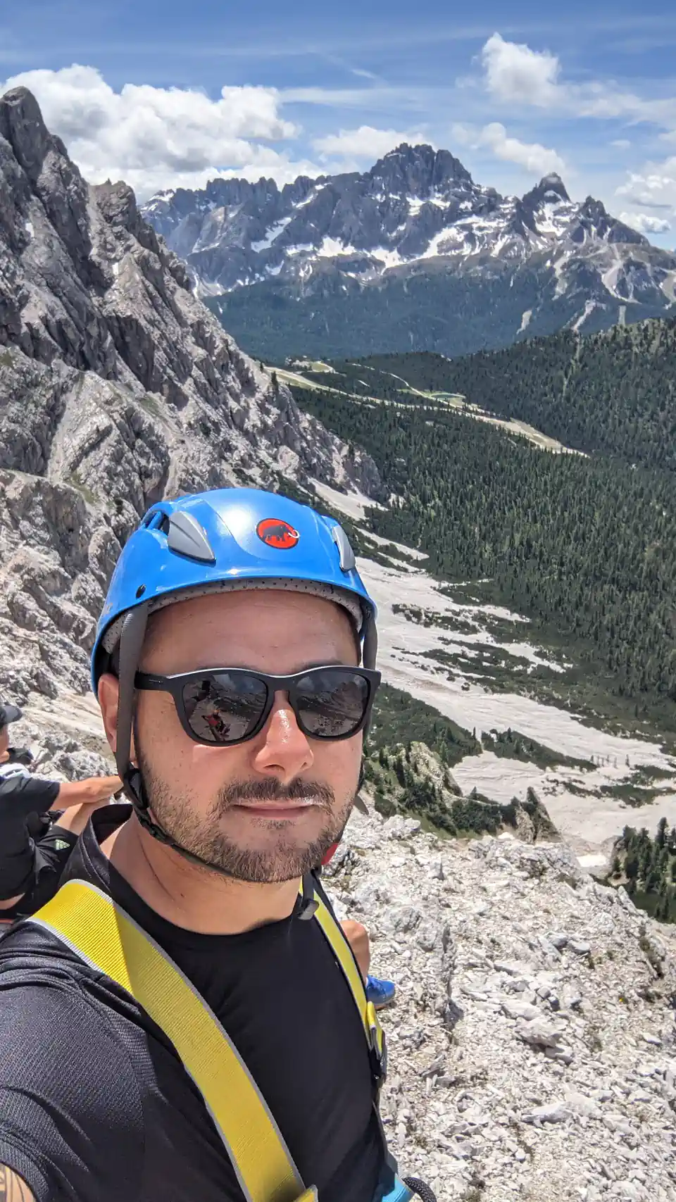 Peter Zošiak se fotí při lezení po via ferratě, s helmou a horolezeckou výbavou v Italii. V pozadí alpská krajina se zelenými údolími a zasněženými horami.