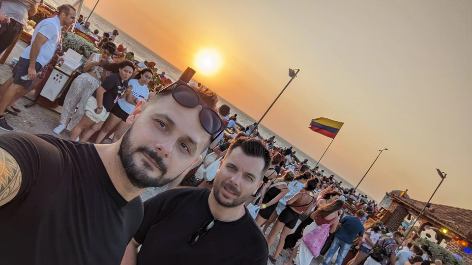 Peter Zošiak s přítelem sledují západ slunce nad mořem v Kolumbii. V pozadí kolumbijská vlajka, pouliční trh a dav turistů si užívají večerní atmosféru.
