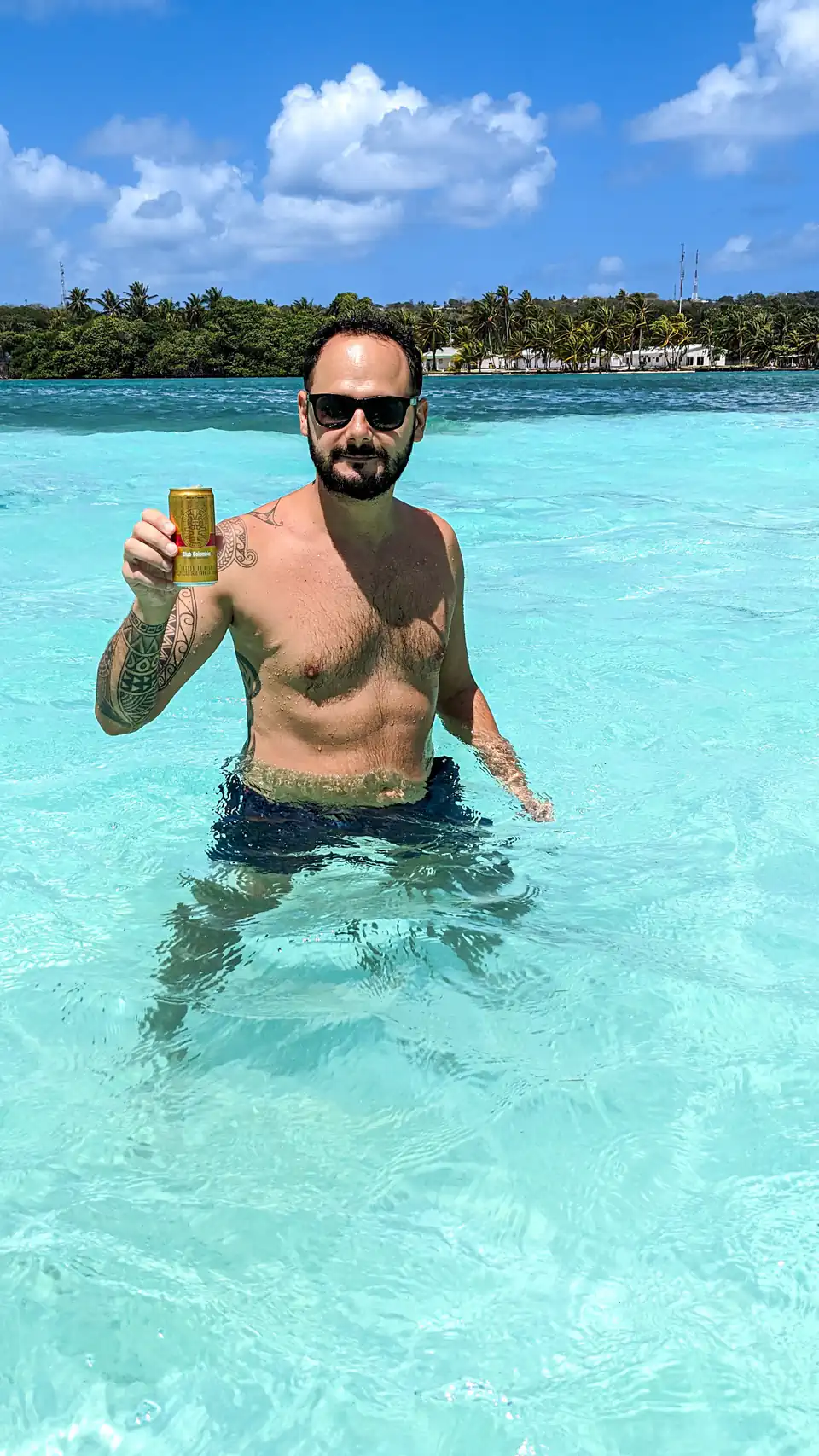 Peter Zošiak stojí v tyrkysově modré vodě na exotické pláži, drží v ruce plechovku piva a nosí sluneční brýle. Za ním palmový ostrov San Andres v Kolumbii a jasná modrá obloha s bílými mraky.