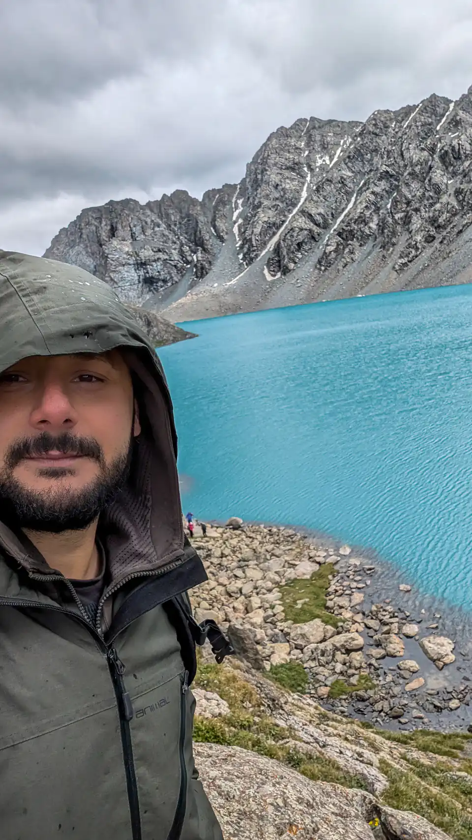 Peter Zošiak se fotí před nádherně modrým horským jezerem Ala-Kul v Kyrgyzstane obklopeným dramatickými skalními štíty. Nosí zelenou outdoorovou bundu s kapucí a nebe je zatažené.